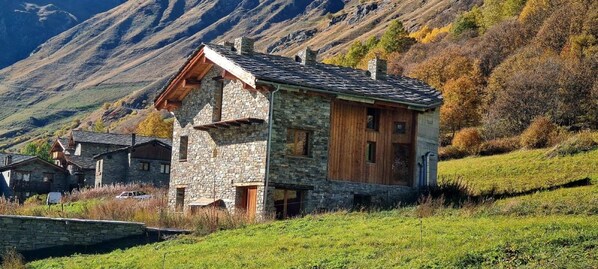 Ett stenhus med en träfasad, omgivet av en grönskande sluttning och berg i bakgrunden.