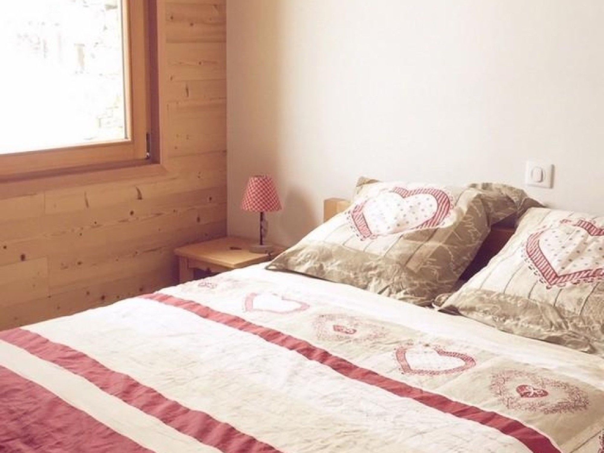 Ein Schlafzimmer mit Holzwand, Fenster, Nachttisch mit Lampe und Bett mit bettenbezogenem Herzmuster.