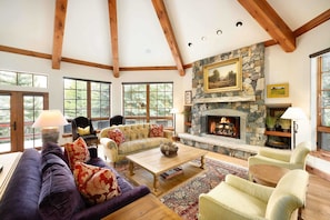 A spacious living room with a stone fireplace, large windows, and a high ceiling with exposed beams. - Spruce Slopeside Ski-in Access (Avon)