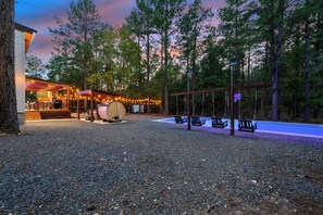 Un espace extérieur aménagé dans un style rustique, avec des bancs en bois et un coin repas abrité, le tout entouré d’arbres et de gravier.