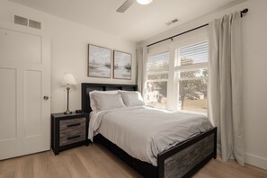 Ein Schlafzimmer mit einem Bett, Nachttischen, einer Lampe, einem Fenster mit Vorhängen und Bildern an der Wand.