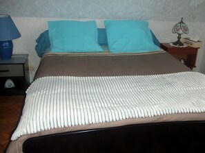 Una cama con una manta a rayas blancas y marrones, dos almohadas azules, una mesita de noche con una lámpara y una caja de pañuelos.