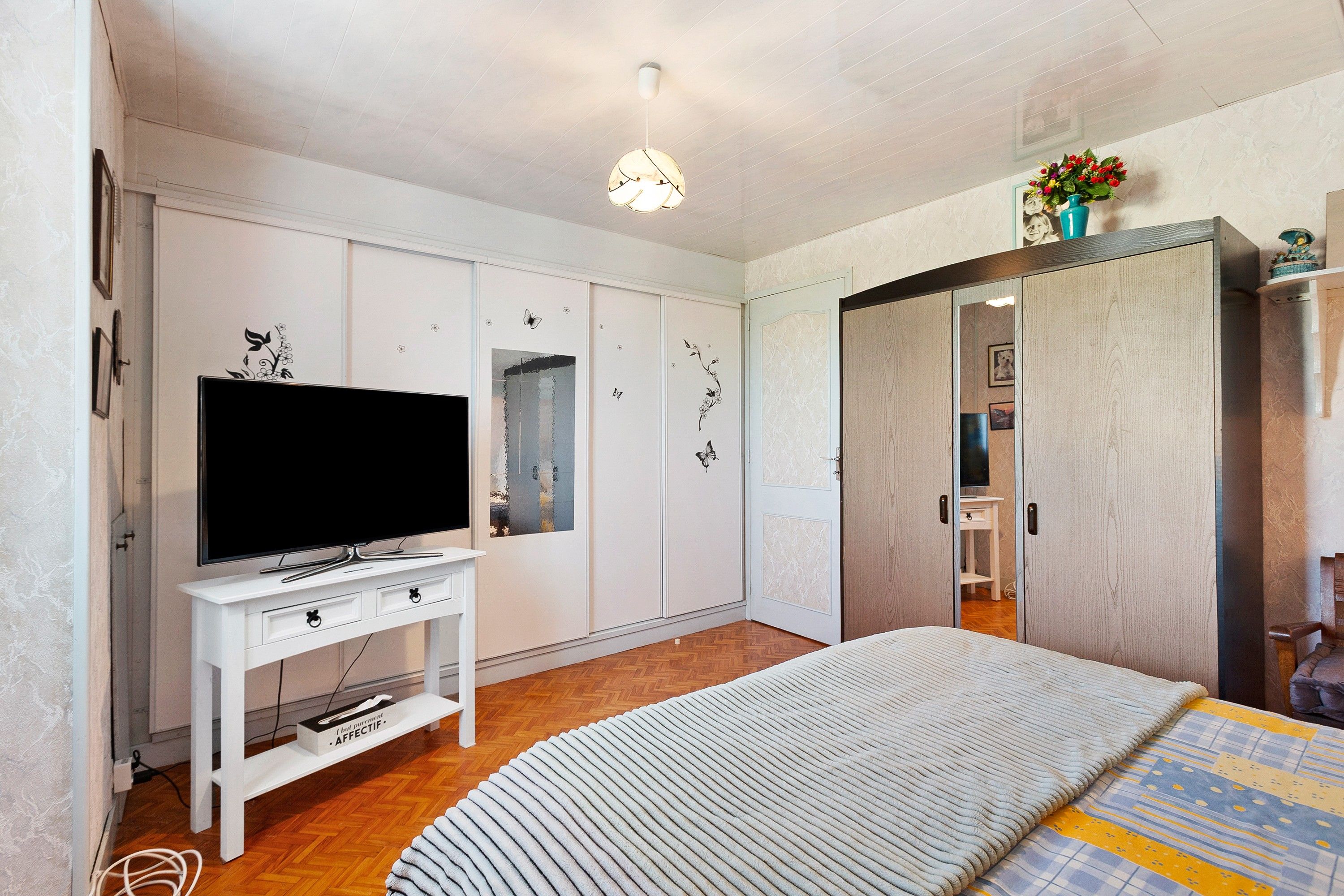 Une chambre à coucher avec un lit, un téléviseur posé sur un meuble blanc, une armoire et un miroir.