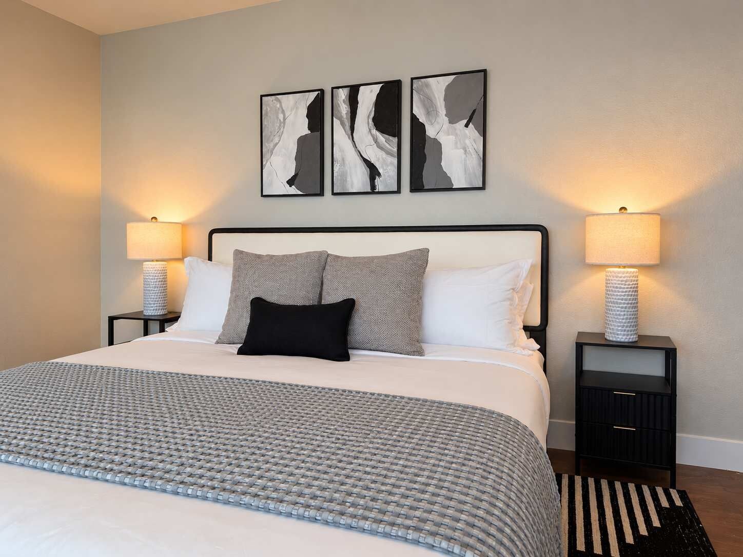A neatly made bed with a patterned bedspread, two bedside lamps, and three abstract wall art pieces above the bed.