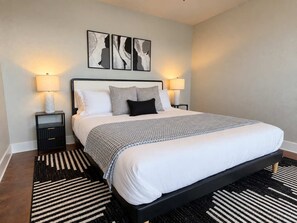 Ein Schlafzimmer mit schwarzem Metallbettgestell, weißer Bettwäsche, zwei Nachttischlampen, einem schwarz-weiß gestreiften Teppich und abstrakter Wandkunst.