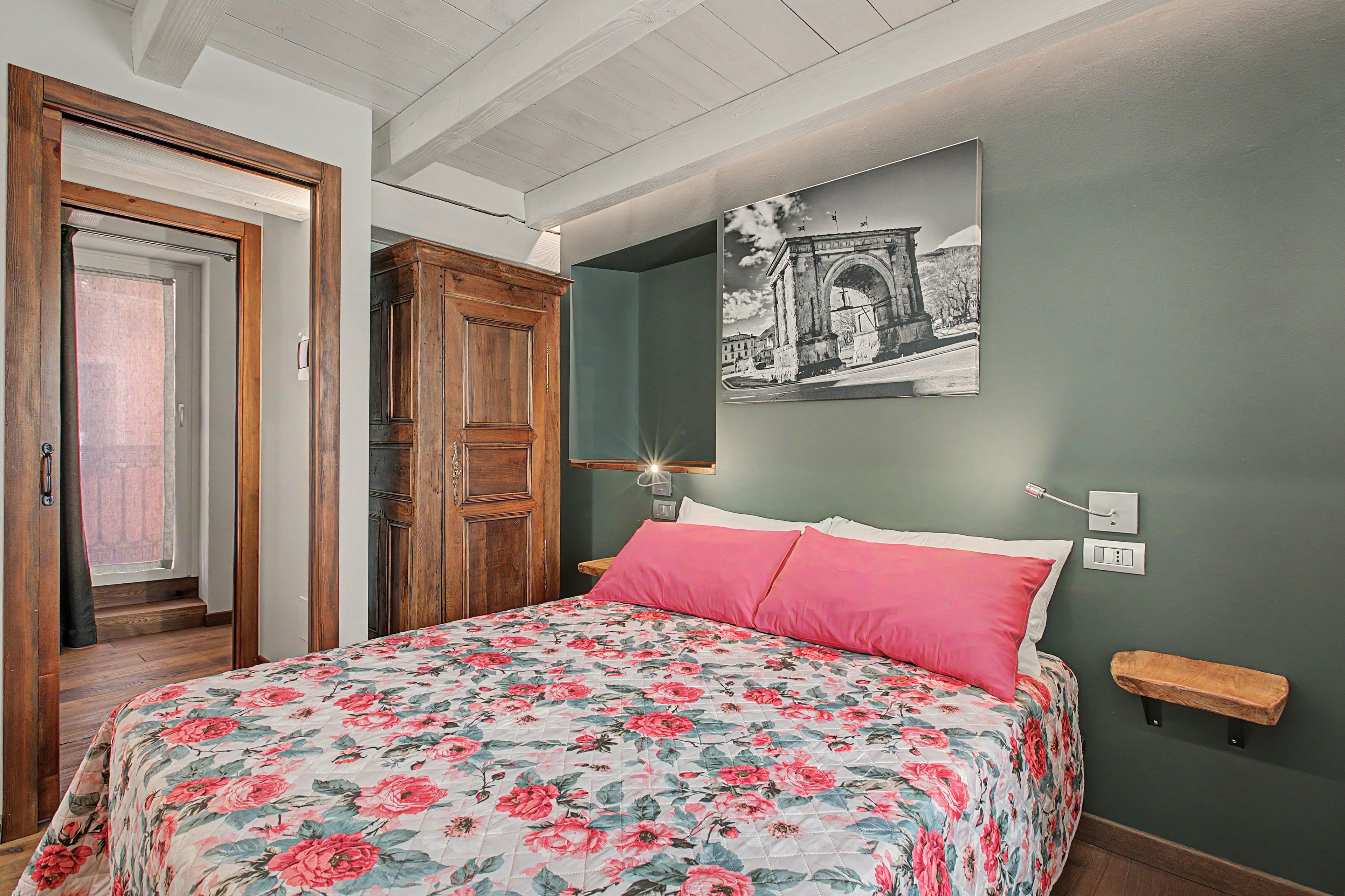 A bedroom with a floral bedspread, a wooden wardrobe, a door with a view, and a wall-mounted light fixture.