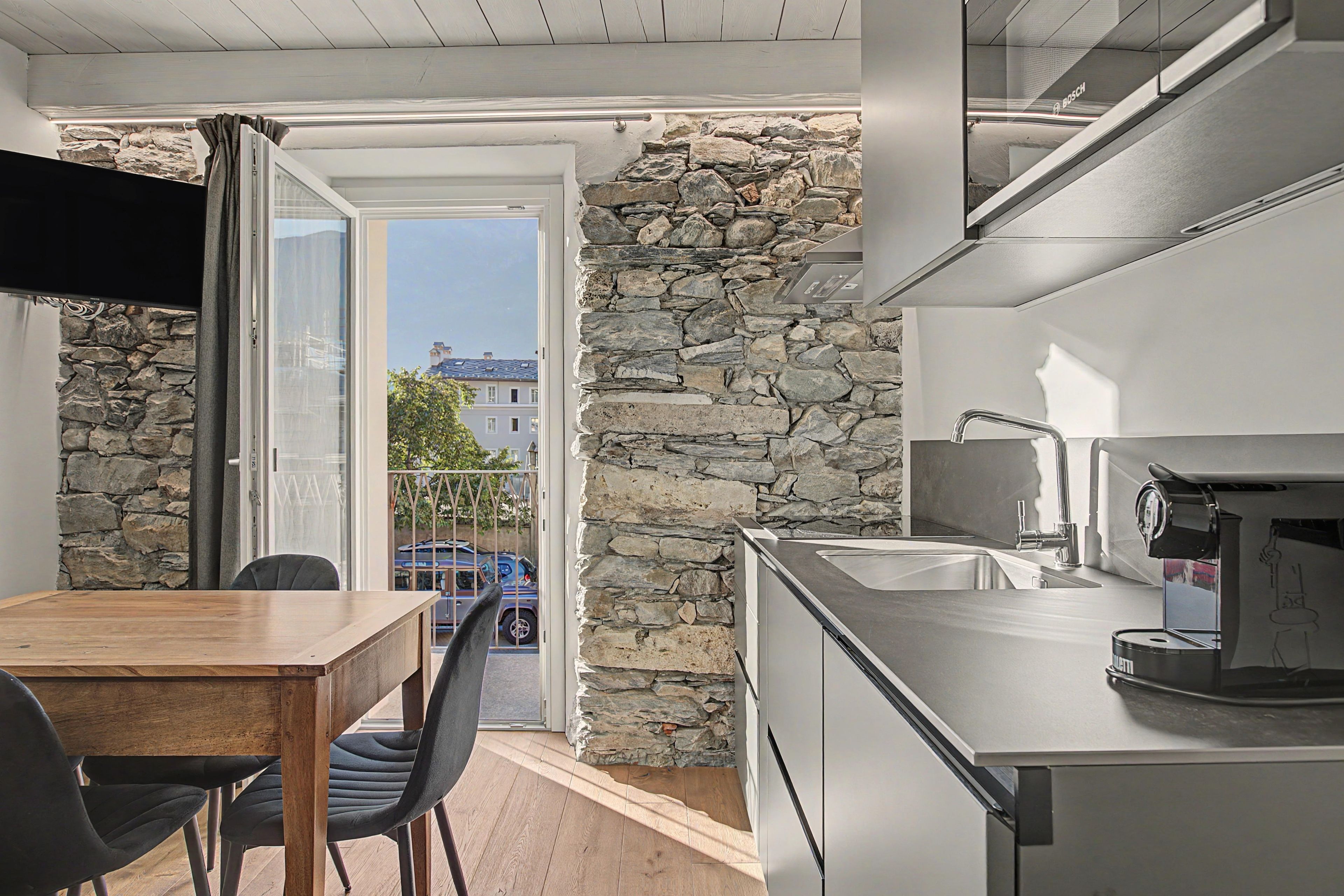 Une cuisine compacte avec un mur en pierre, un coin repas avec une table et des chaises en bois, et une vue sur l’extérieur.