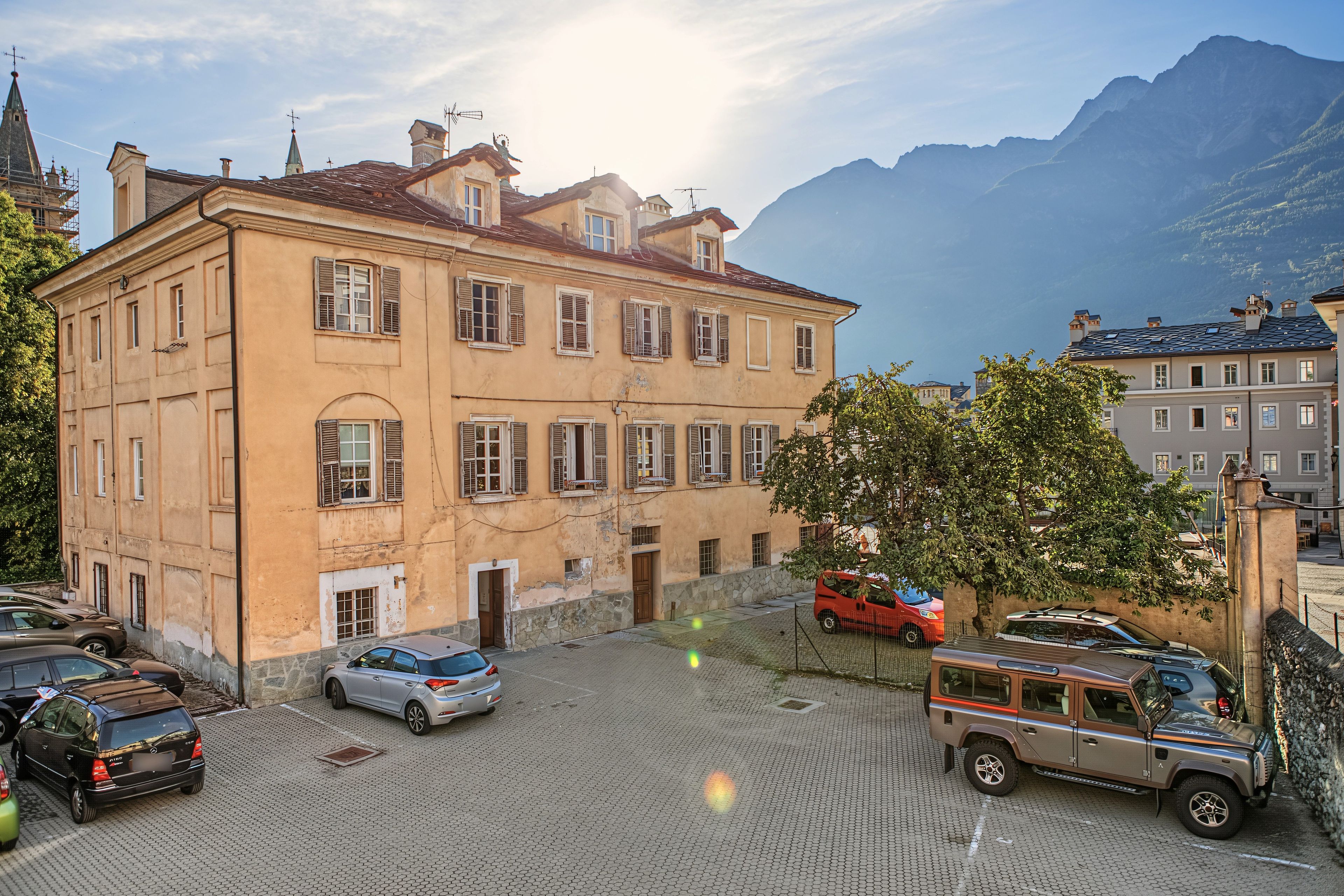 Un edificio histórico con patio, coches estacionados y montañas al fondo.