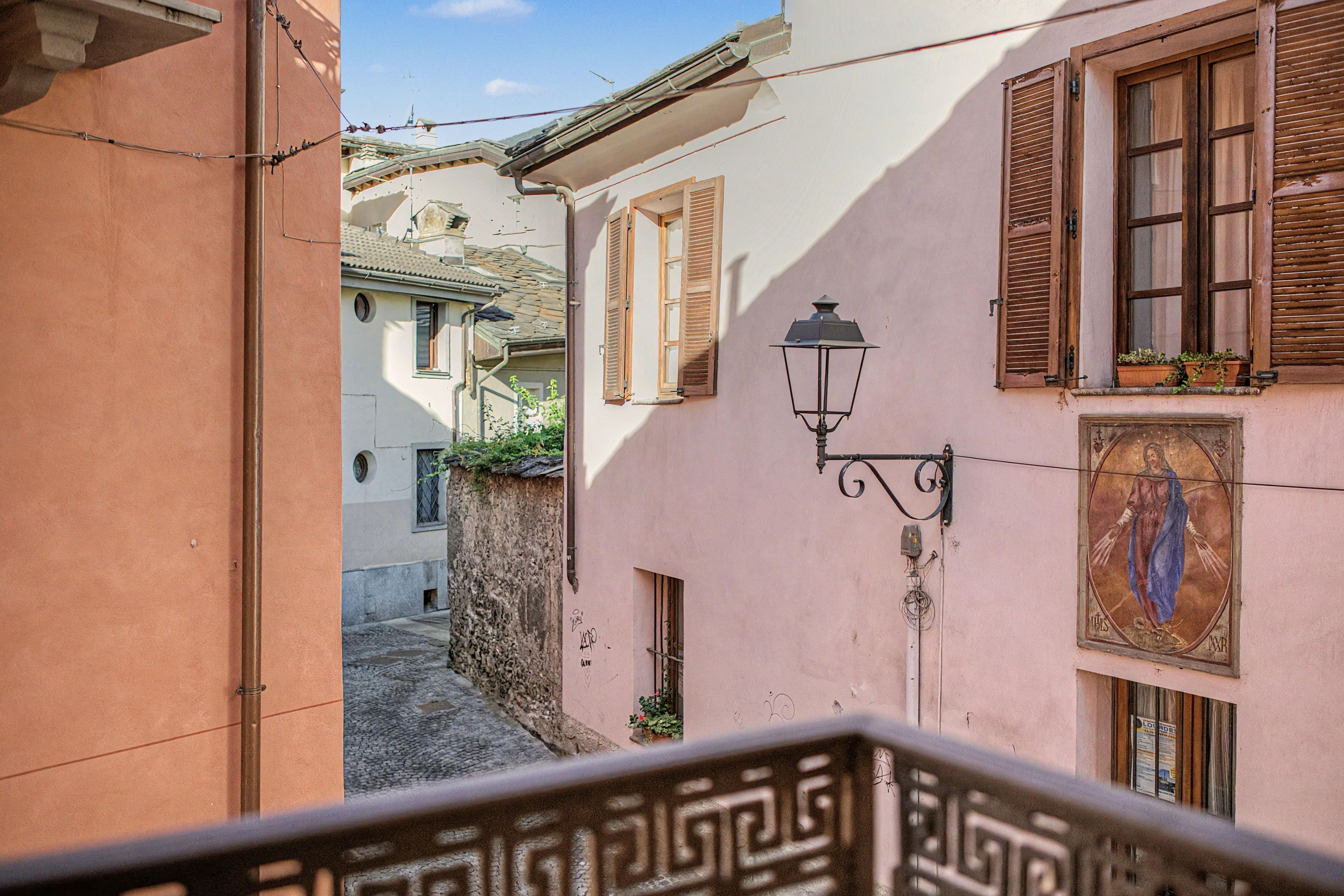 A narrow street with buildings on either side, a street lamp, and a small balcony with a decorative railing.