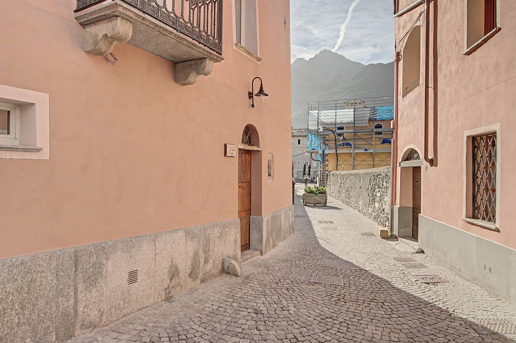 Una stretta strada acciottolata affiancata da edifici con balconi e una montagna sullo sfondo.