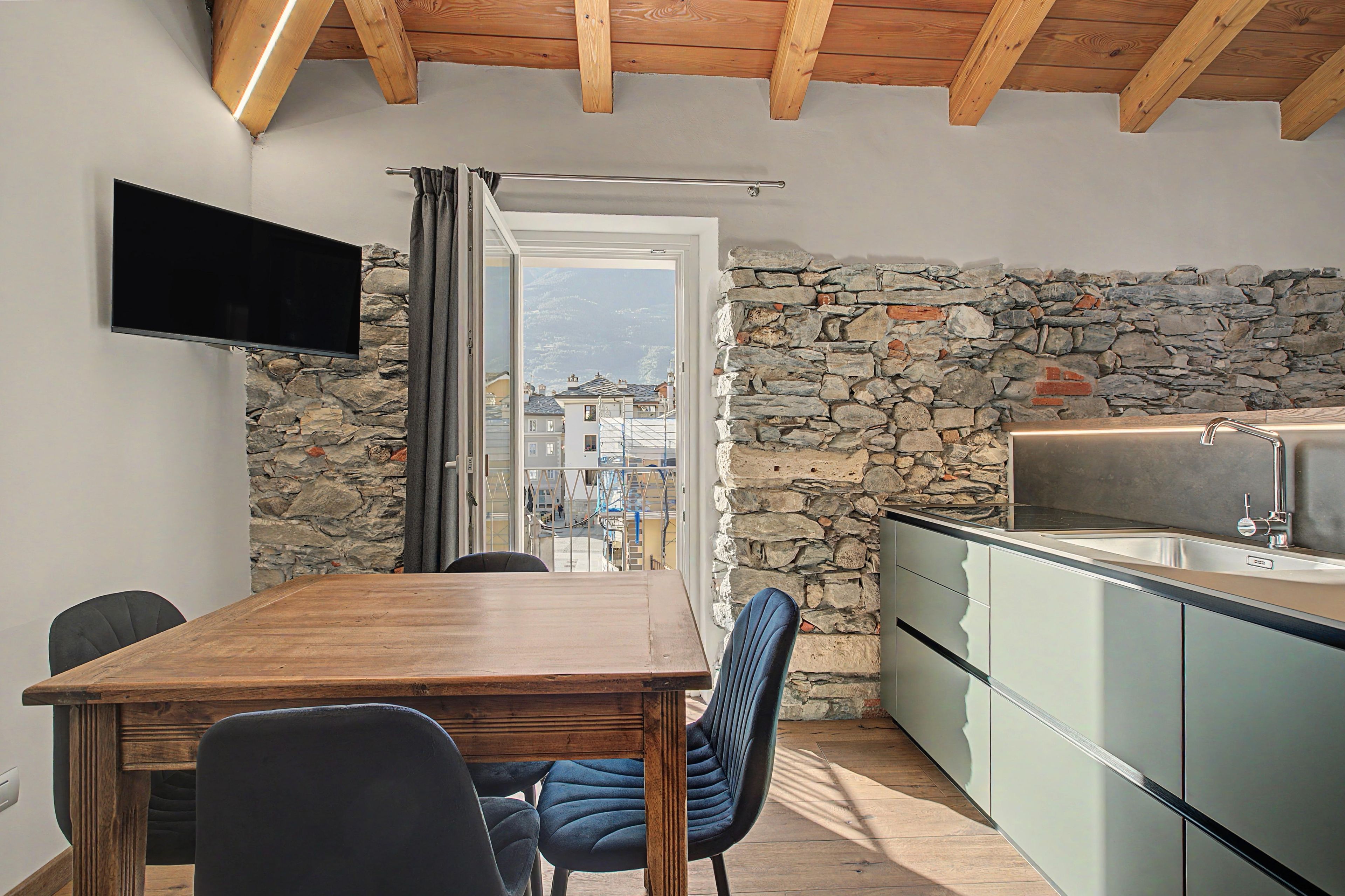 A dining area with a wooden table and chairs, a stone wall, a kitchen with a sink, and a TV mounted on the wall.