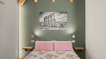 Un dormitorio con una cama, dos mesitas de noche, una lámpara de pared e imagen enmarcada.
