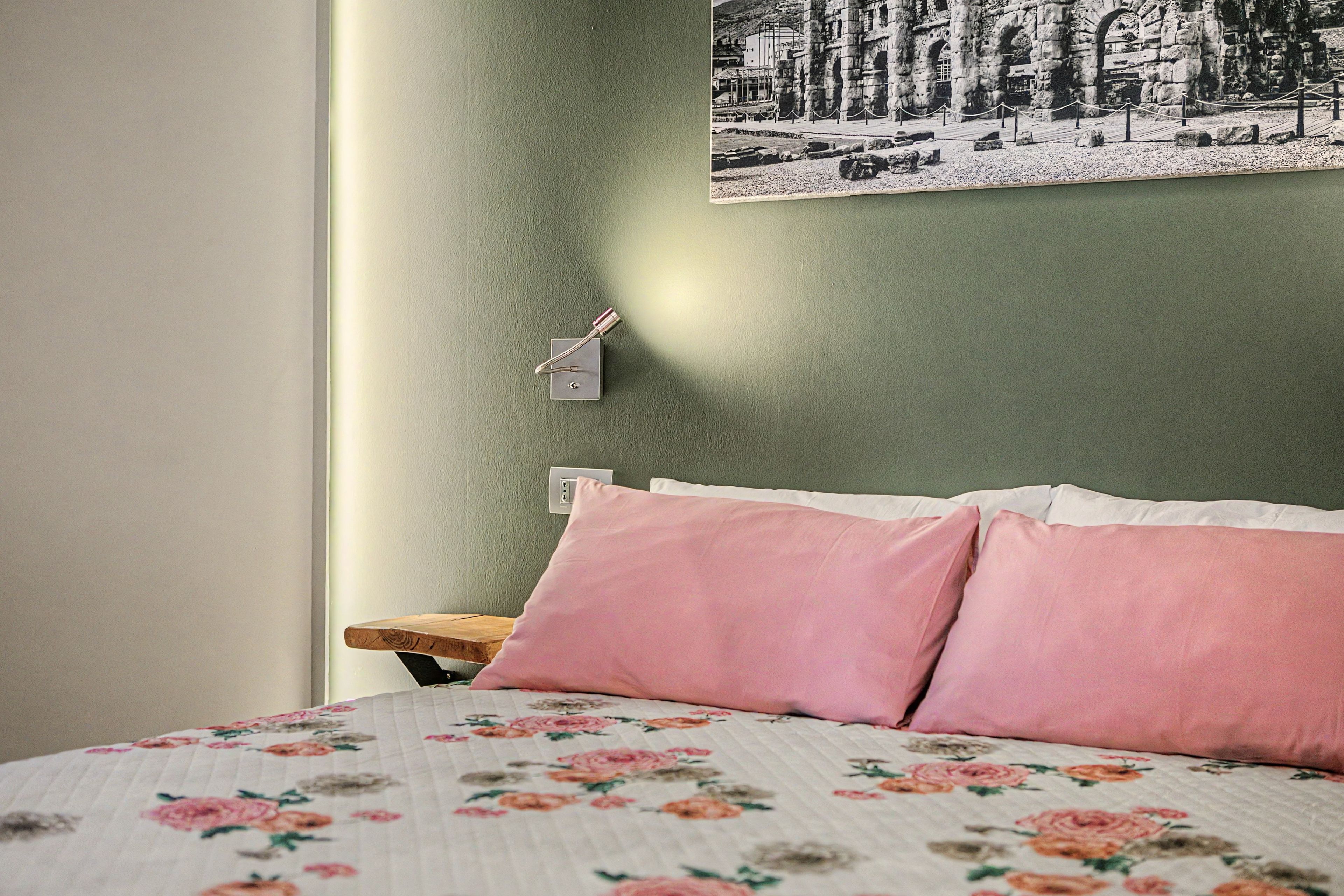 Una camera da letto con un letto, cuscini, un comodino, un interruttore a parete e un quadro incorniciato.