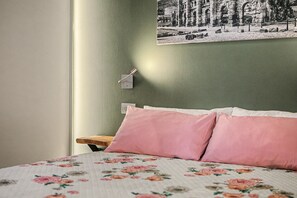 Une chambre à coucher avec un lit, des oreillers, une table de chevet, un interrupteur fixé au mur et un tableau encadré accroché au mur.