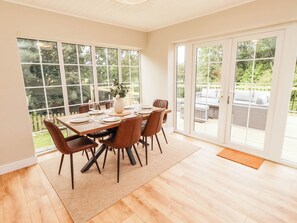 A dining area with a large table set for six, surrounded by chairs, with a view of the outdoors through large windows. - Hedley Dene Cottage (Morpeth)