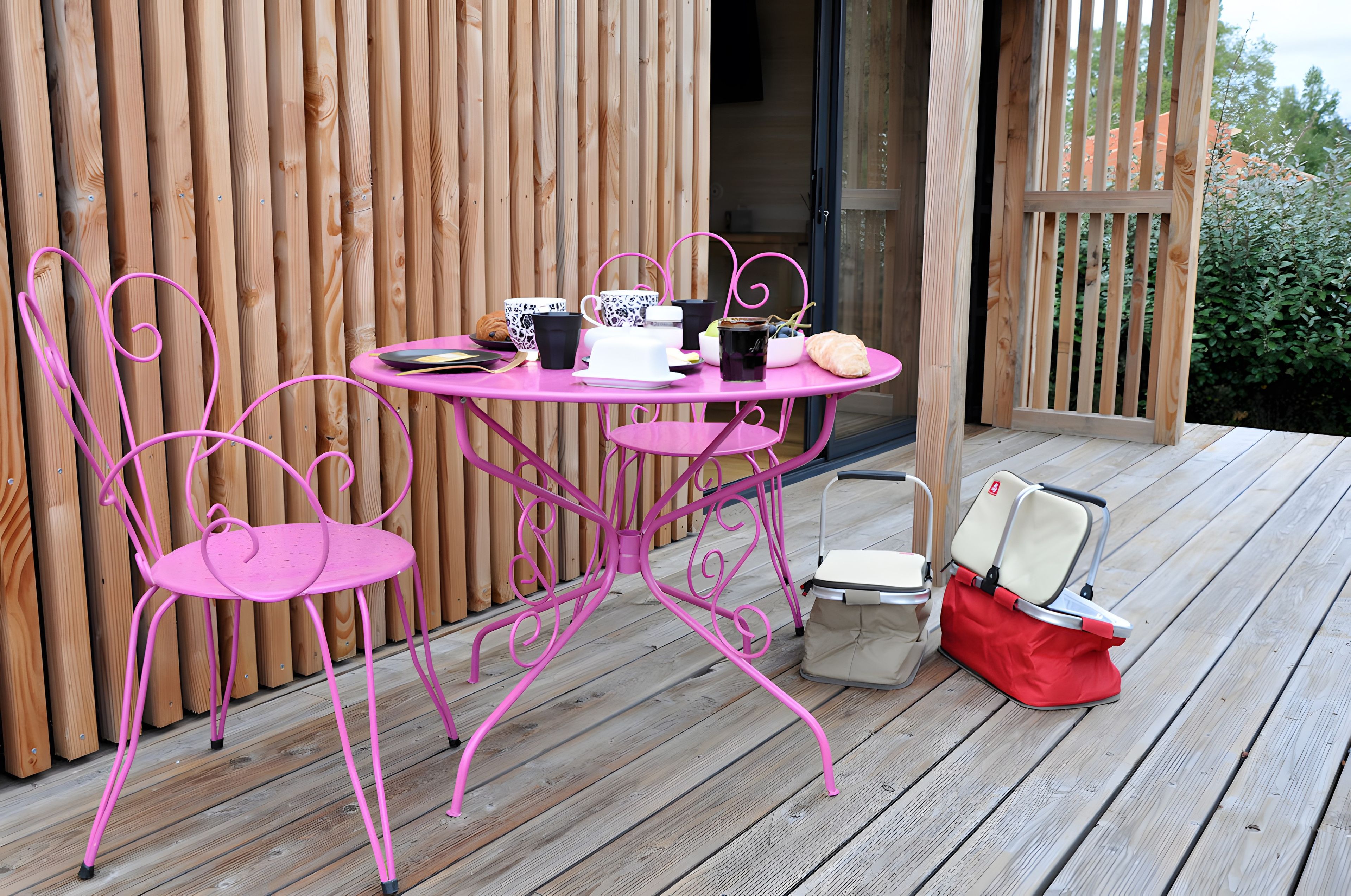 Eine Holzterrasse mit einem pinkfarbenen Metalltisch und -stühlen, ein kleiner Tisch mit Tasse und Croissant sowie eine rote Tasche am Boden.