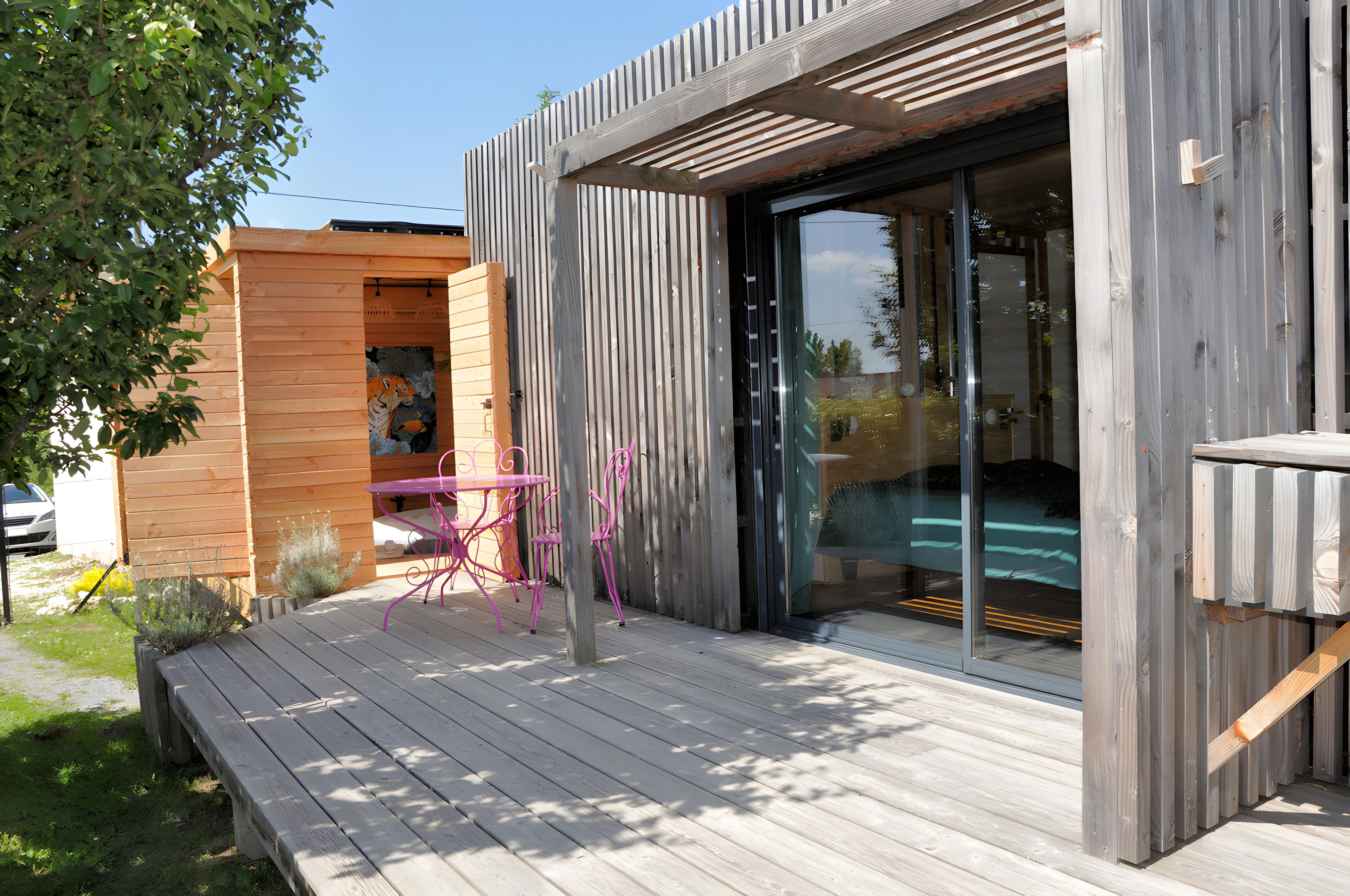 A modern outdoor patio with a wooden deck, a glass door entrance, and a pink metal table and chairs.