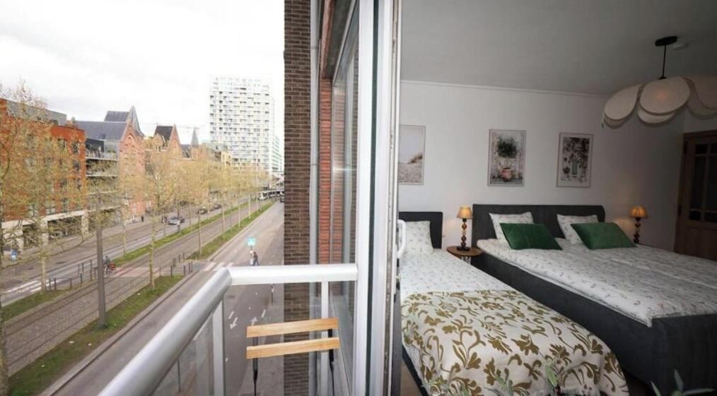 Een slaapkamer met een groot bed, een raam met uitzicht op een straat en gebouwen, en een balkon met een hekwerk.