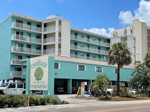 Un hôtel à plusieurs étages doté de balcons, une enseigne indiquant « The Chateaux Condo Miniums » au 19440 Gulf Blvd, et un espace de stationnement agrémenté d’un palmier.