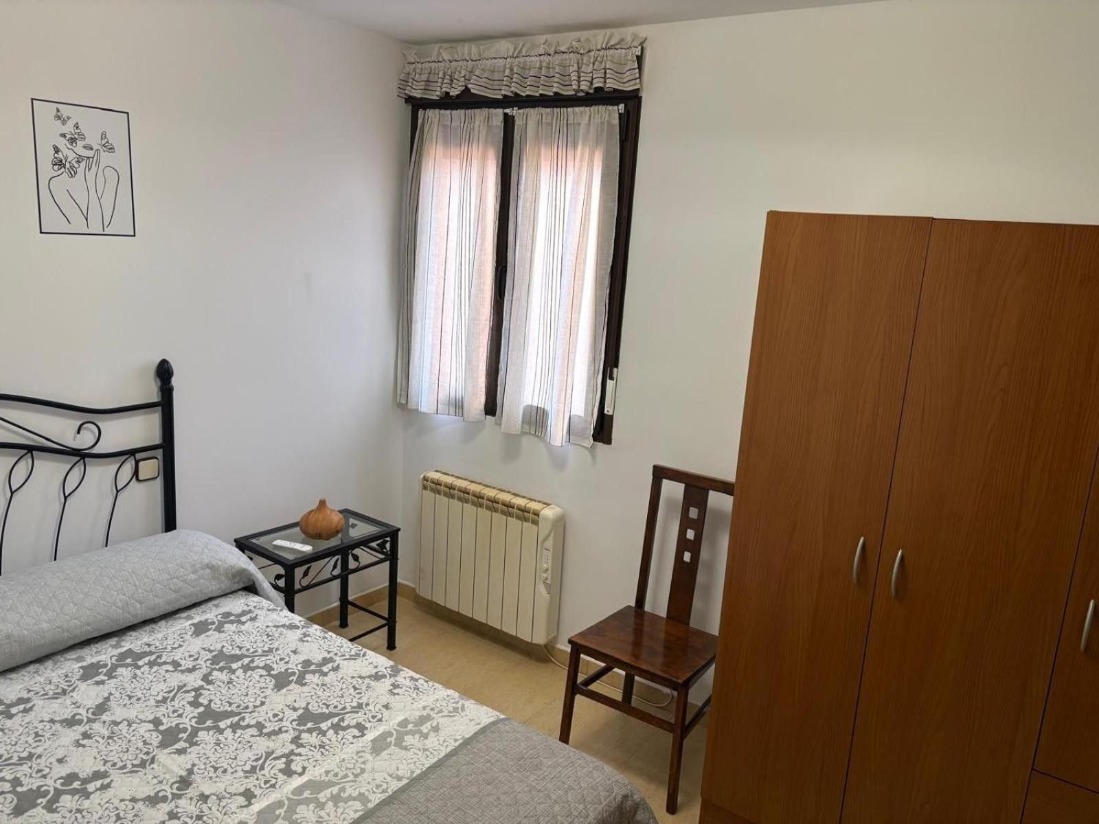 A bedroom with a bed, a wooden chair, a radiator, a window with curtains, and a wardrobe.