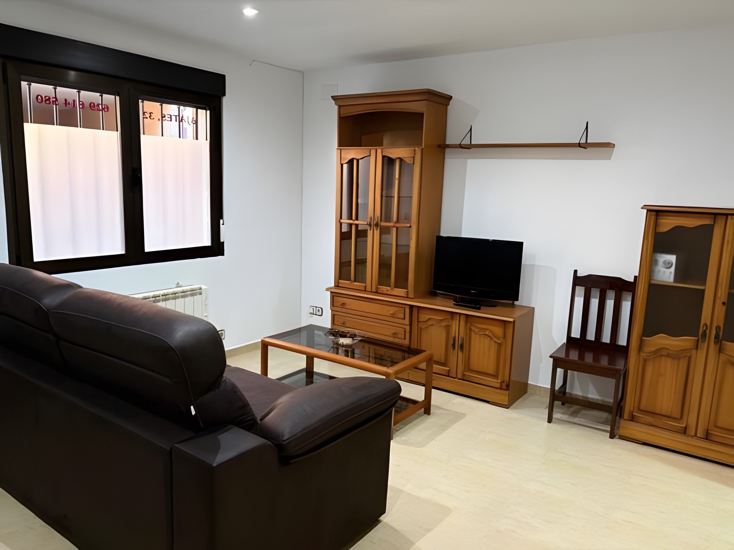 A living room with a leather sofa, a glass coffee table, wooden cabinets, a television, and a window with blinds.