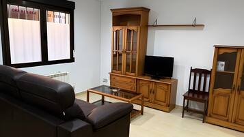 A living room with a leather sofa, a glass coffee table, wooden cabinets, a television, and a window with blinds.