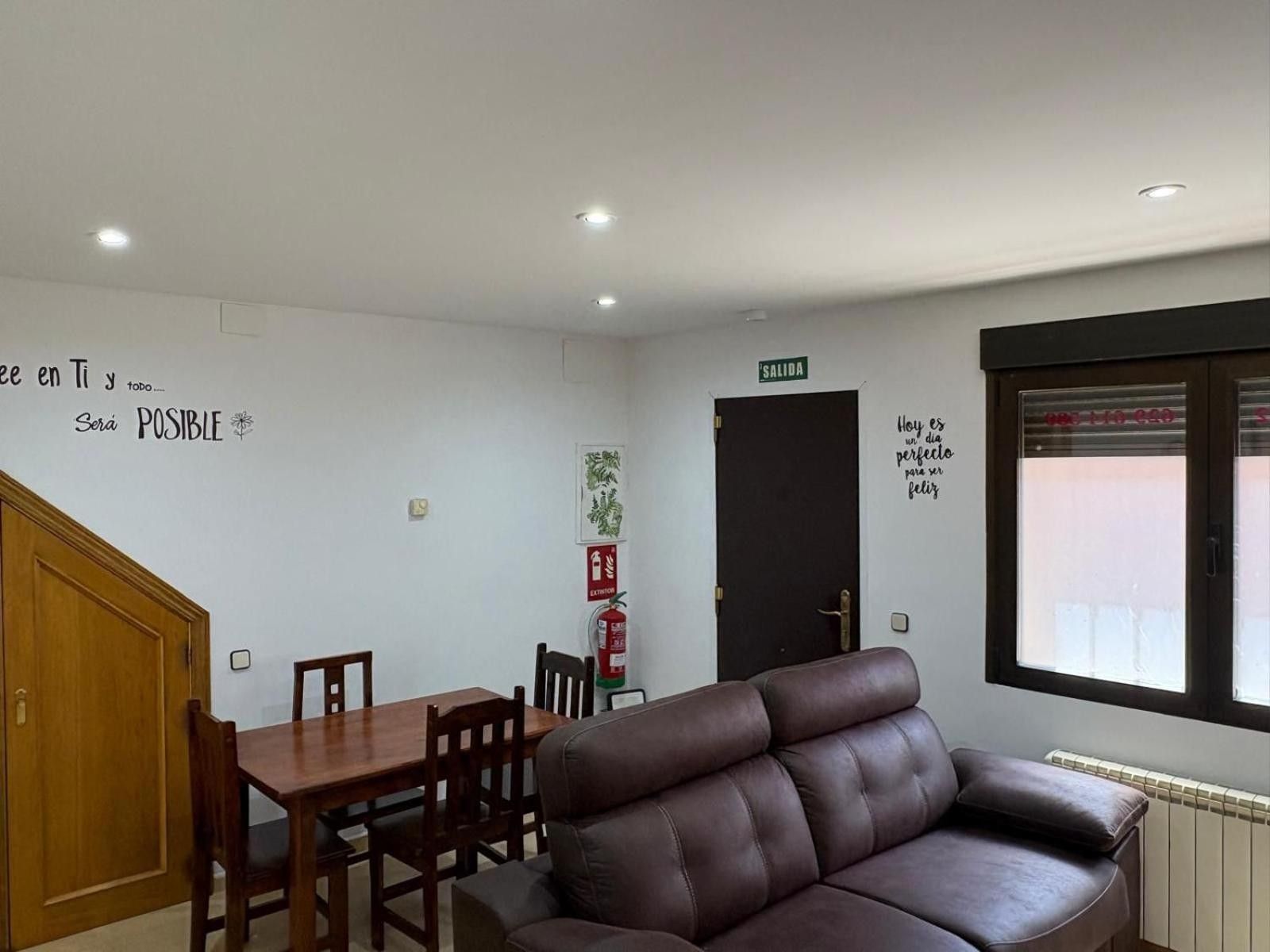 A room with a dining table, chairs, a sofa, a door with a sign, and a window with a view.