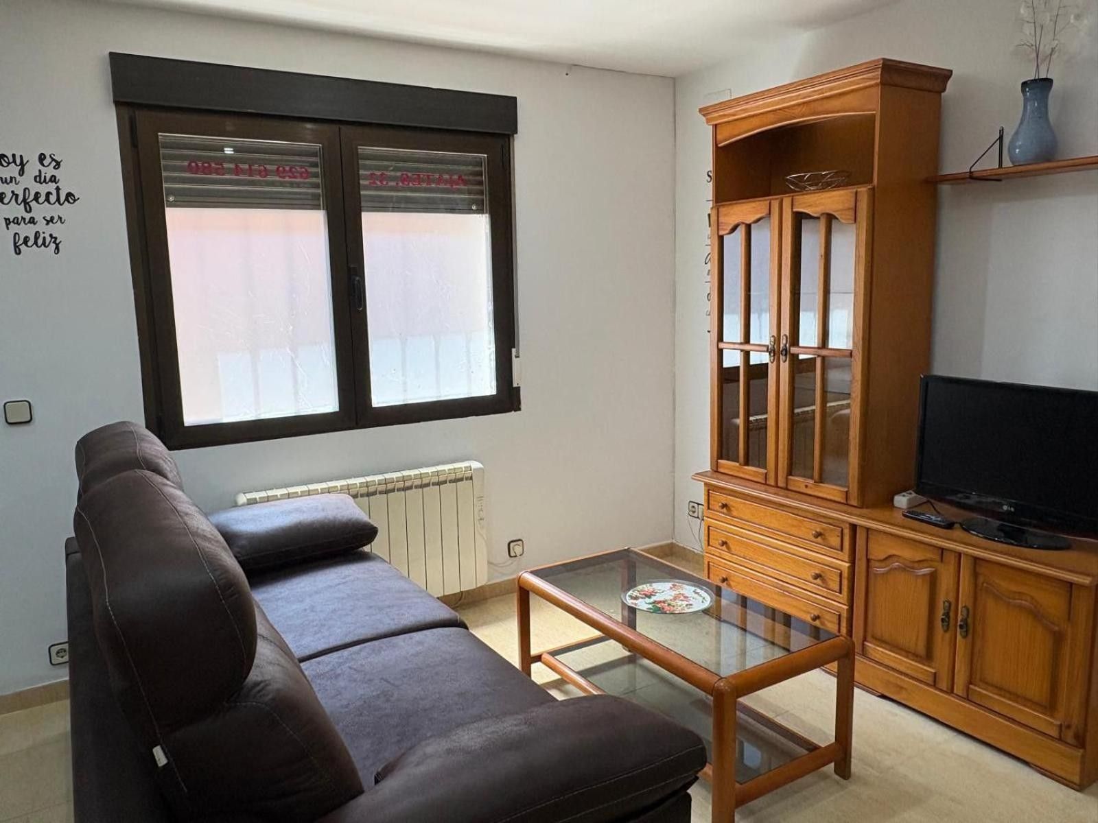 Una sala de estar con un sofá marrón, una mesa de centro de vidrio, un mueble de madera y un televisor.