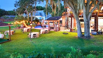 An outdoor patio area with tables and chairs, surrounded by trees and a pool.