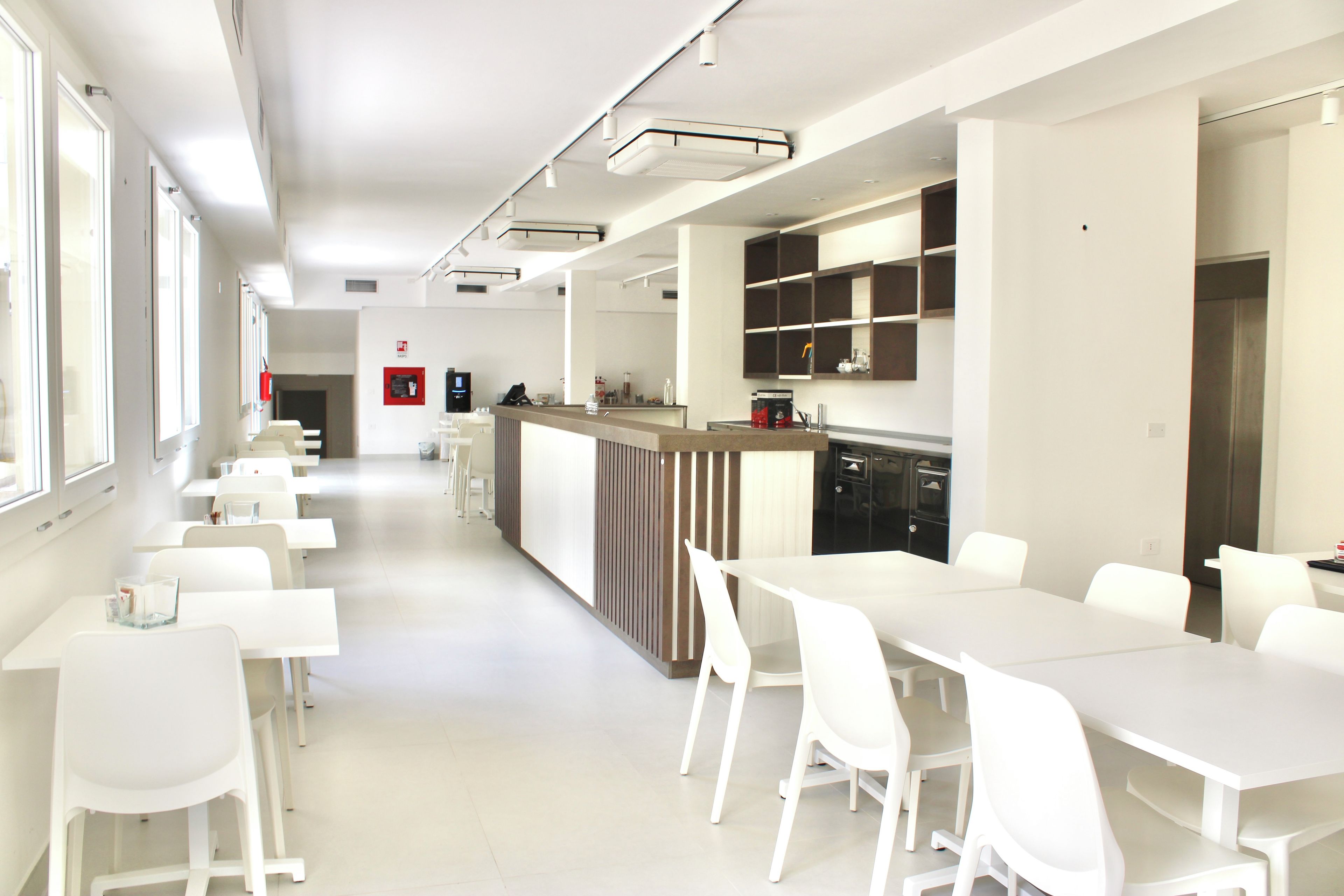 Un espace repas moderne et bien éclairé, avec des tables et des chaises blanches, un coin cuisine doté d’un comptoir et de placards, et un détecteur d’incendie fixé au mur.