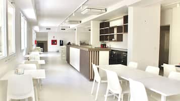 Un espace repas moderne et bien éclairé, avec des tables et des chaises blanches, un coin cuisine doté d’un comptoir et de placards, et un détecteur d’incendie fixé au mur.
