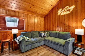 A cozy cabin-style room with a wooden ceiling and walls, a green upholstered sectional sofa, two side tables with lamps, and a window with a view of trees. - 5 Primary En-Suites on First Floor, Hot Tub, Sauna (Wintergreen Resort)