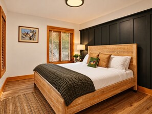 Quarto com cama de madeira, janela com persianas, luminária na parede e quadro emoldurado.