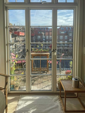 A room with a large sliding glass door leading to an outdoor area with a wooden table and chairs, a small rug, and a metal trash can. - Two-floor flat with balcony in Vesterbro (København V)