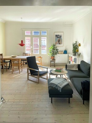 A living room with a sofa, armchair, and ottoman, a dining area with chairs and a table, and a bookshelf with books and decorative items. - Two-floor flat with balcony in Vesterbro (København V)