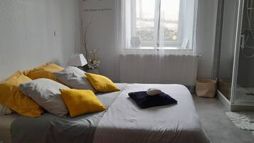 Une chambre à coucher avec un lit, des oreillers, une table de chevet, une lampe, un vase avec des fleurs, une fenêtre et un espace douche.