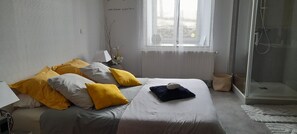 Een slaapkamer met een bed, kussens, een nachtkastje, een lamp, een vaas met bloemen, een raam en een douchehoek.