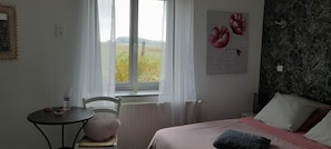 A bedroom with a bed, a chair, a table with a water bottle, and a window with a view of greenery.