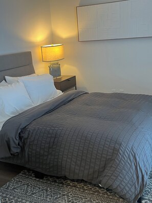 Ein ordentlich bezogenes Bett mit einer grauen Tagesdecke, einer Nachttischlampe und einem kleinen Nachttisch mit einer Lampe.