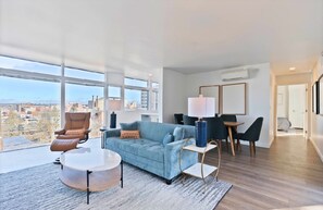 Ein modernes Wohnzimmer mit Sofa, Sessel und Couchtisch, ein Essbereich mit Stühlen und große Fenster mit Blick auf die Stadt.