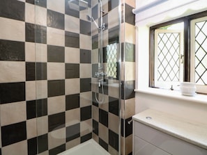A bathroom with a checkered tile wall, a glass shower enclosure, a window with a lattice design, and a sink with a white countertop. - Tudor Lodge (Belper)