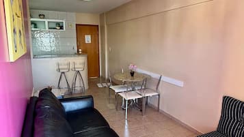 Un saló amb un sofà de cuir negre, una zona de menjador amb una taula rodona i cadires, i una cuina amb rajoles blanques i una porta de fusta.