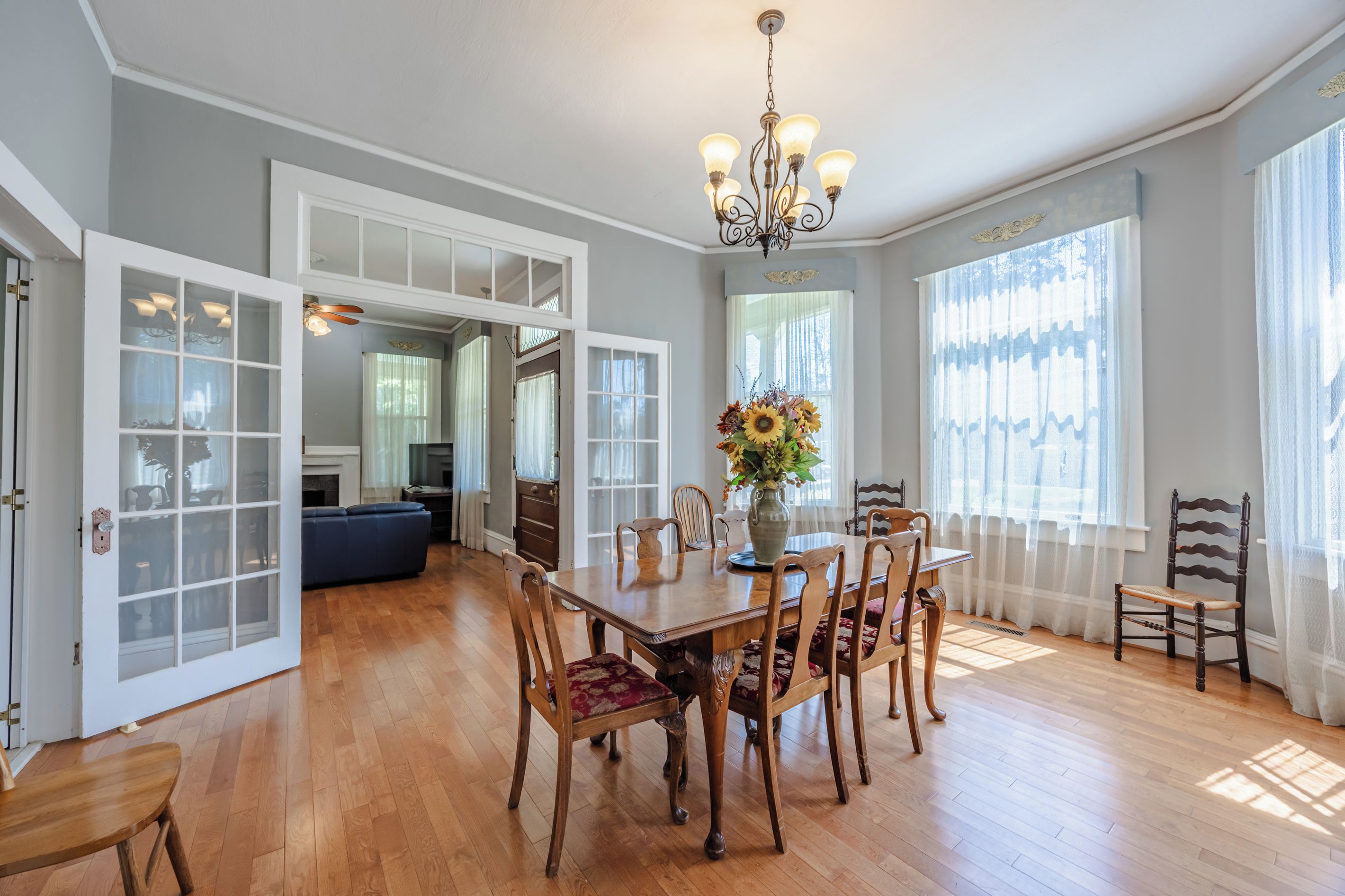 A dining area with a wooden table, chairs, and a vase of flowers. There are large windows with curtains and a chandelier above.