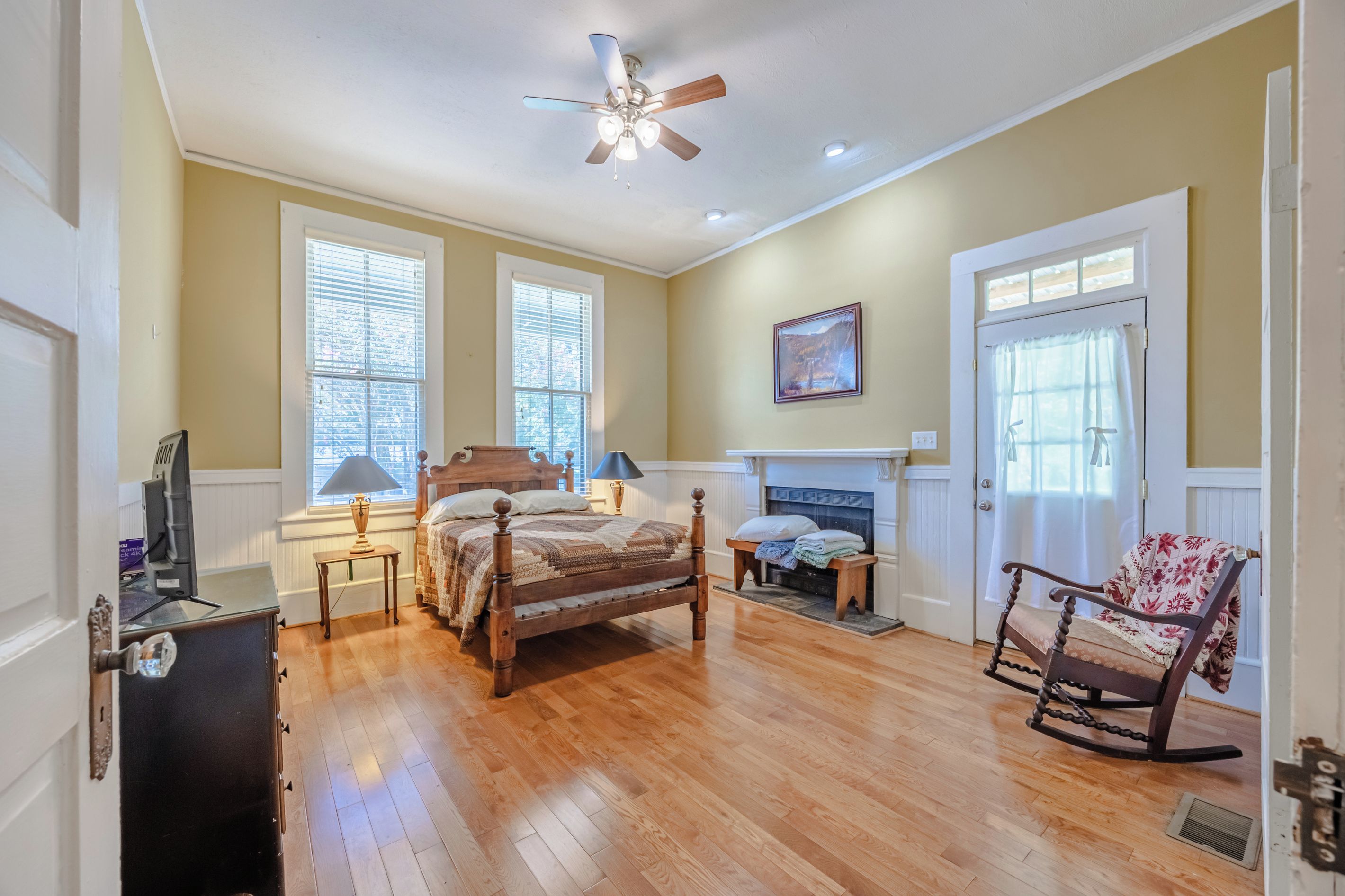 A bedroom with a wooden bed, a fireplace, a rocking chair, and a ceiling fan.