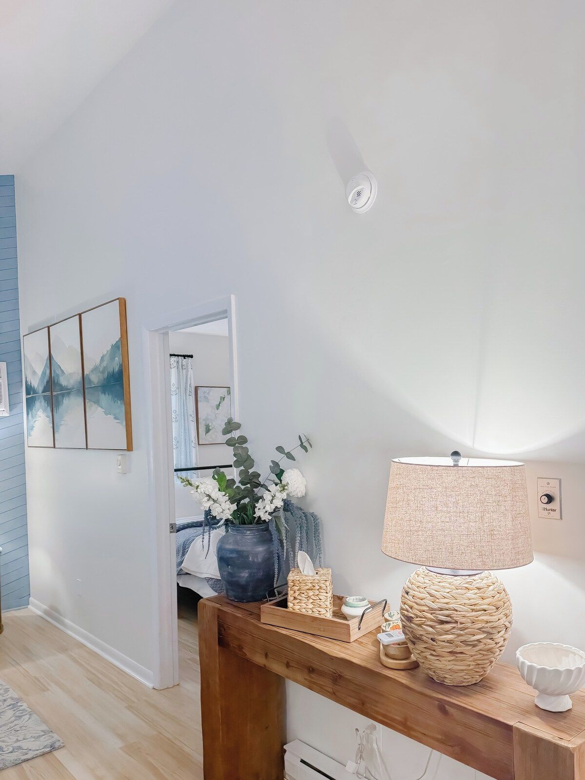 A room with a wooden console table, a lamp, a vase with flowers, and a framed picture on the wall.