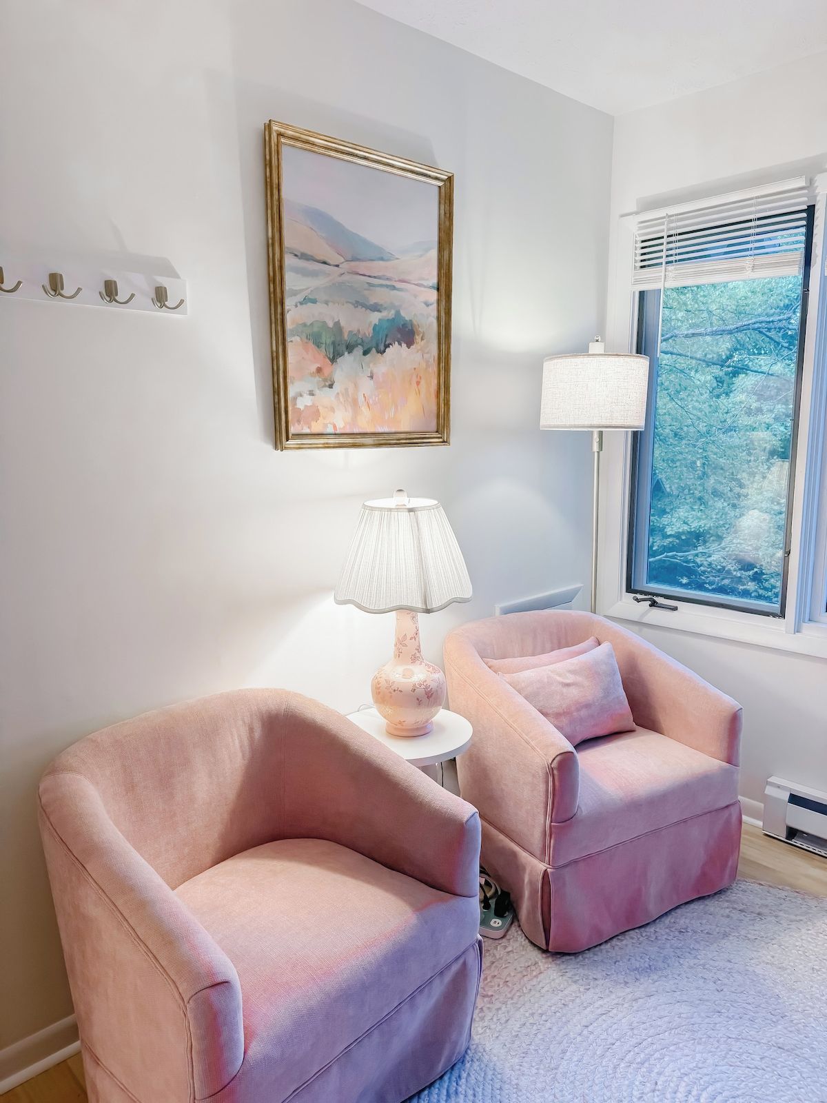 A room with two pink armchairs, a lamp, a painting, and a window with blinds.