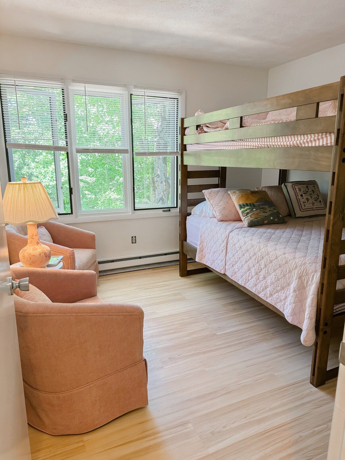 A bedroom with a bunk bed, a cushioned chair, a bedside table with a lamp, and a window with blinds.