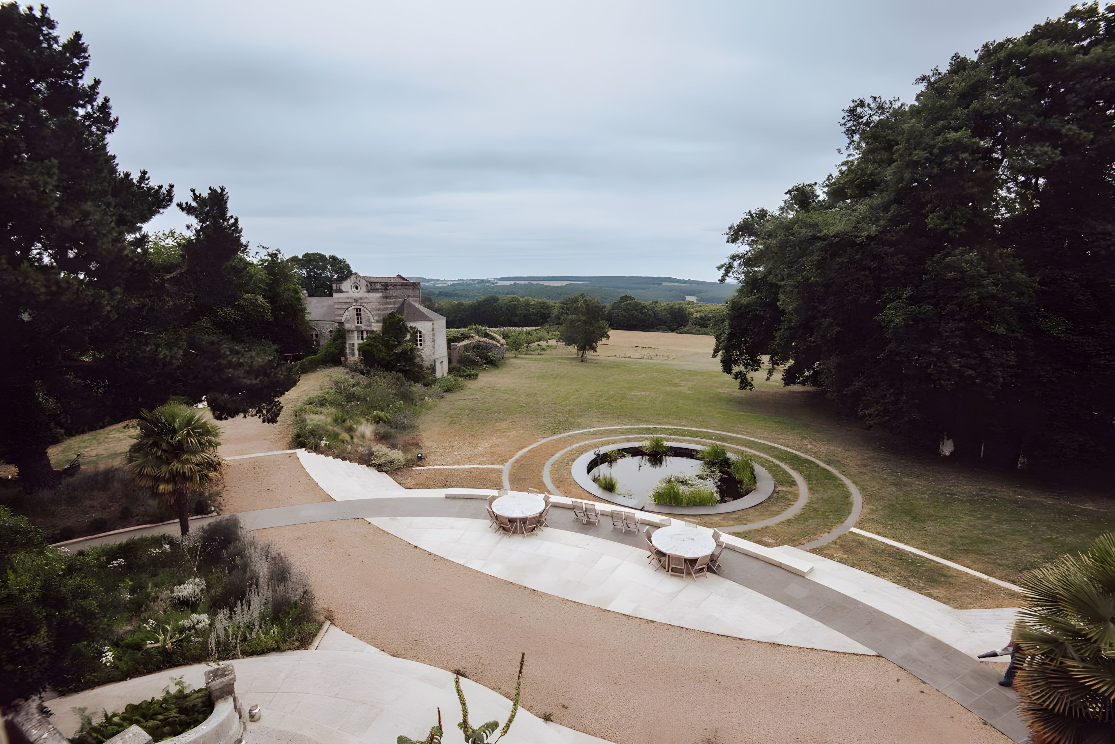 A landscaped garden with a circular water feature, seating areas, and a view of a distant building.