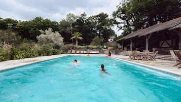 A swimming pool with two people in the water, surrounded by greenery and a covered area with chairs.