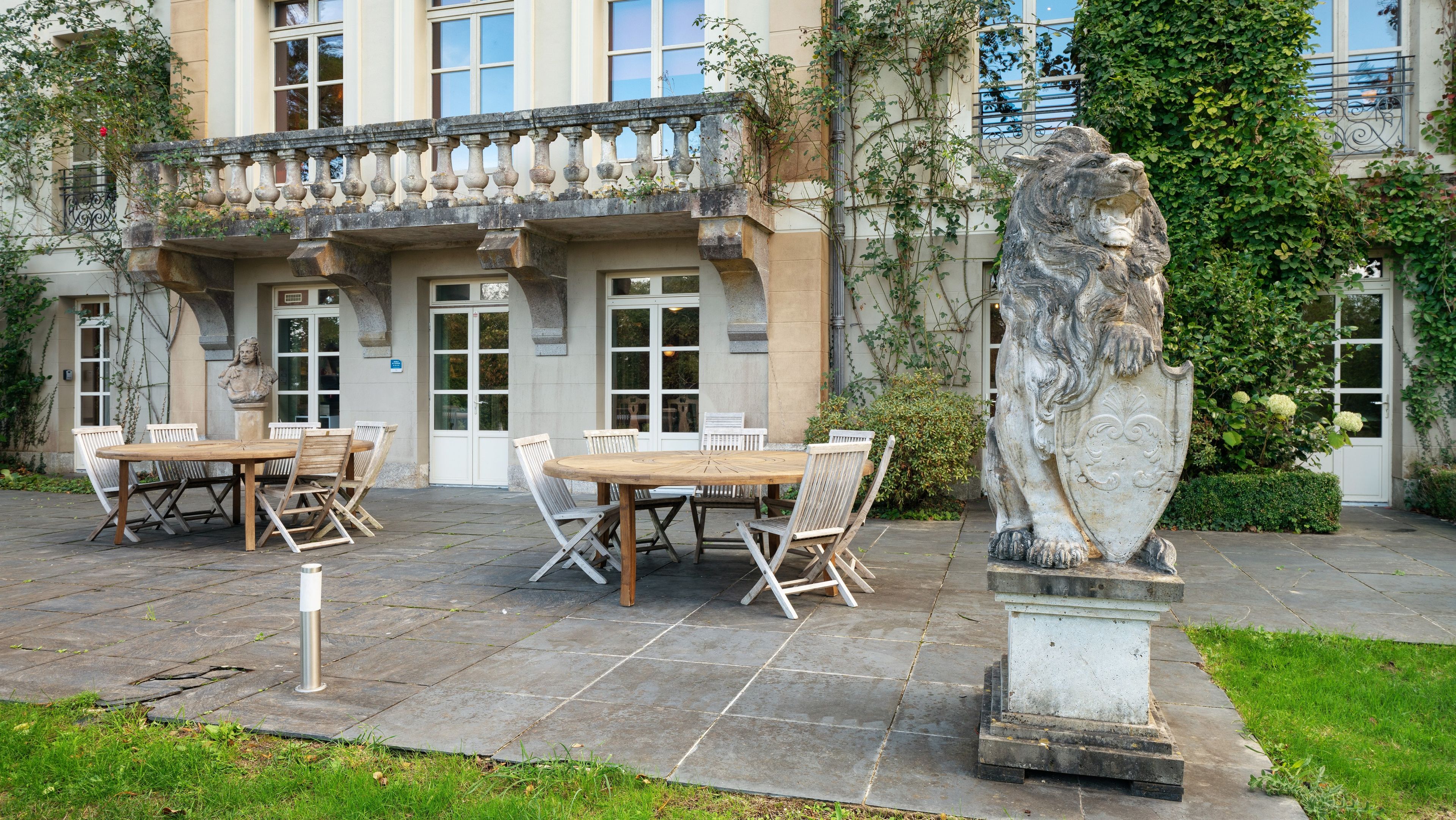 Un espace extérieur aménagé avec une table et des chaises en pierre, une statue et un bâtiment doté de grandes fenêtres et d’un balcon.